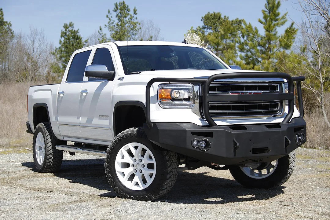 Fab Fours Premium Winch front bumper with full guard on a white GMC Sierra 1500, front passenger-side three-quarter view outdoors, showing full guard hoop, winch plate, round fog light pods