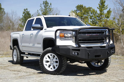 Fab Fours Premium Winch front bumper with full guard on a white GMC Sierra 1500, front passenger-side three-quarter view outdoors, showing full guard hoop, winch plate, round fog light pods