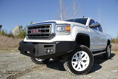 Fab Fours Premium Winch front bumper on a white GMC Sierra 1500, front driver-side three-quarter view outdoors, showing winch plate, round fog light pods, squared end caps
