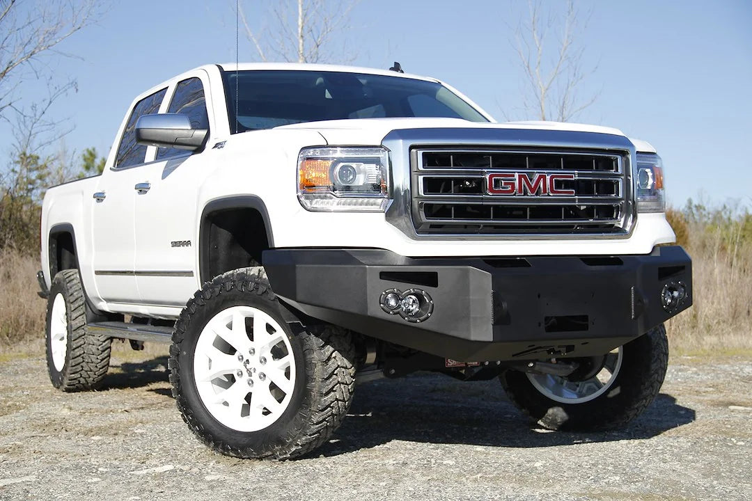Fab Fours Premium Winch front bumper on a white GMC Sierra 1500, front passenger-side three-quarter view outdoors, showing winch plate, round fog light pods, squared end caps