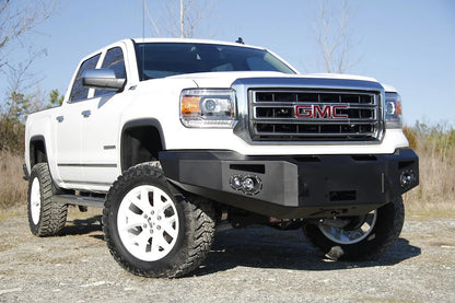 Fab Fours Premium Winch front bumper on a white GMC Sierra 1500, front passenger-side three-quarter view outdoors, showing winch plate, round fog light pods, squared end caps