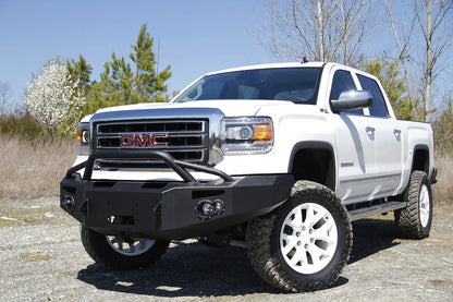 Fab Fours Premium Winch front bumper with pre-runner guard on a white GMC Sierra 1500, front driver-side three-quarter view outdoors, showing pre-runner hoop, winch plate, round fog light pods