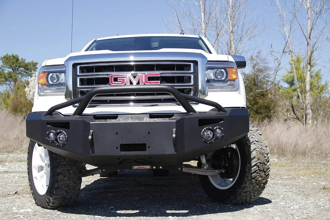 Fab Fours Premium Winch front bumper with pre-runner guard on a white GMC Sierra 1500, straight-on front view outdoors, showing pre-runner hoop, winch plate, round fog light pods