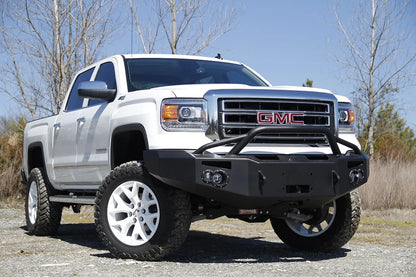 Fab Fours Premium Winch front bumper with pre-runner guard on a white GMC Sierra 1500, front passenger-side three-quarter view outdoors, showing pre-runner hoop, winch plate, round fog light pods