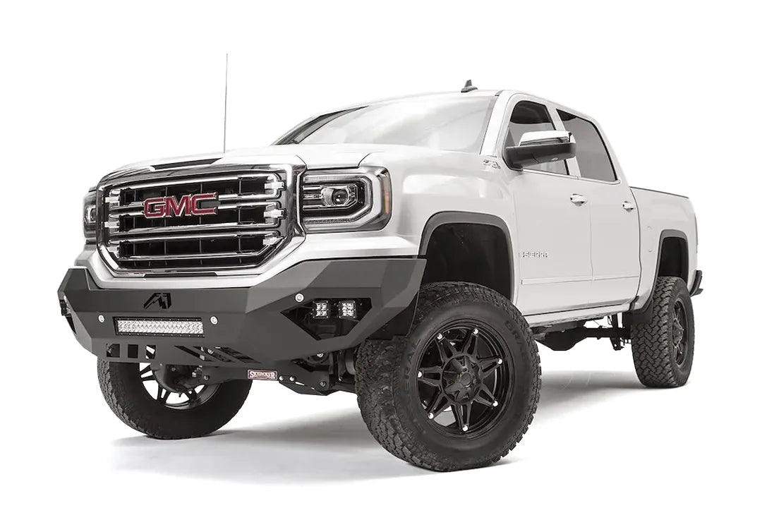 Fab Fours Vengeance front bumper on a white GMC Sierra 1500, driver-side front three-quarter view, showing the angular bumper face, pod light cutouts and lower light bar mount