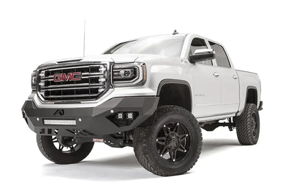 Fab Fours Vengeance front bumper on a white GMC Sierra 1500, driver-side front three-quarter view, showing the angular bumper face, pod light cutouts and lower light bar mount