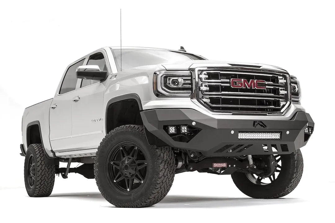 Fab Fours Vengeance front bumper on a white GMC Sierra 1500, passenger-side front three-quarter view, showing the angular bumper face, pod light cutouts and lower light bar mount