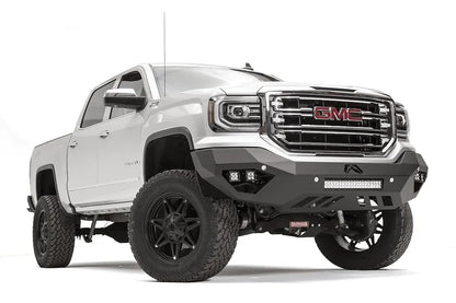 Fab Fours Vengeance front bumper on a white GMC Sierra 1500, passenger-side front three-quarter view, showing the angular bumper face, pod light cutouts and lower light bar mount