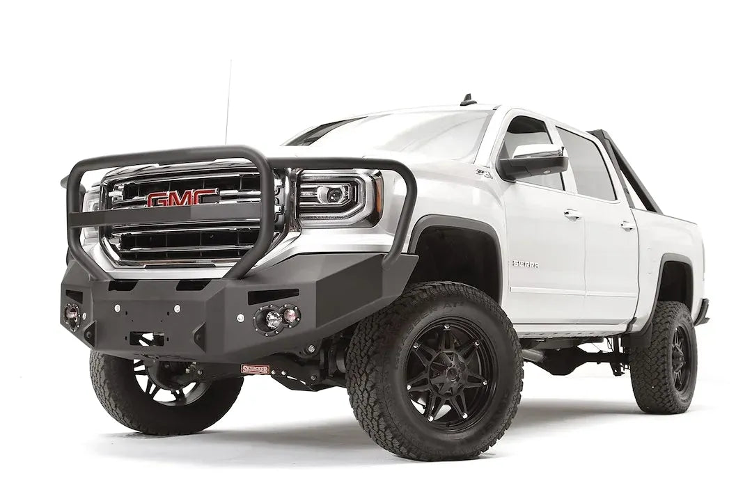Fab Fours Premium Winch front bumper with full guard on a white GMC Sierra 1500, driver-side front three-quarter view, showing the full grille guard, winch fairlead opening and round fog lights