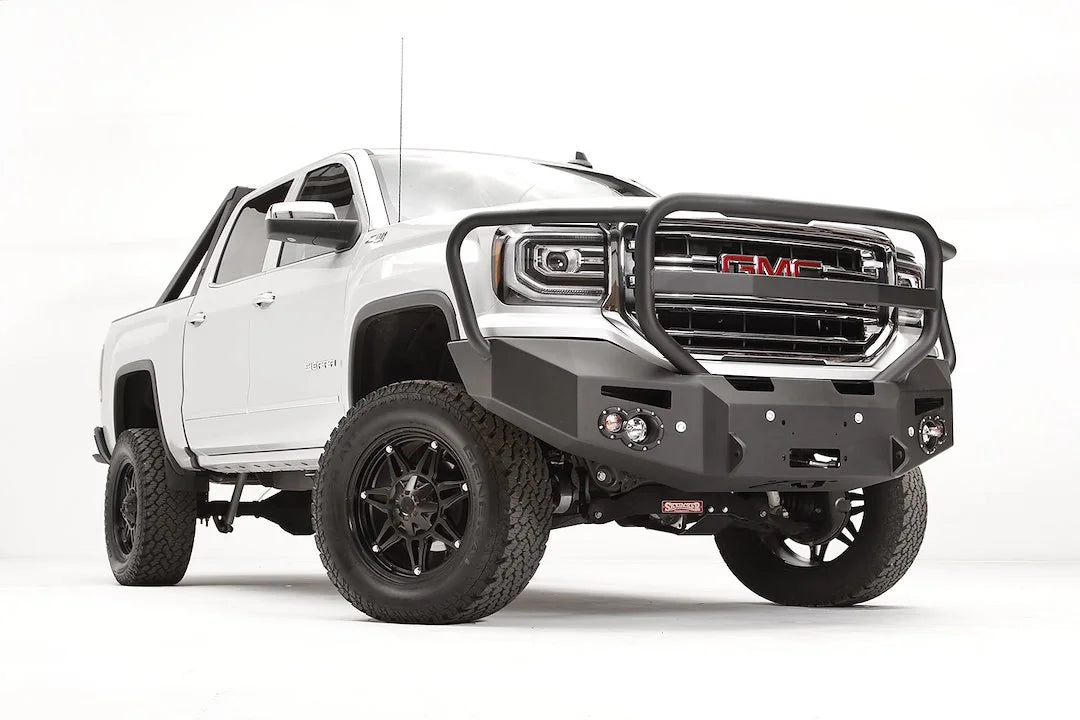 Fab Fours Premium Winch front bumper with full guard on a white GMC Sierra 1500, passenger-side front three-quarter view, showing the full grille guard, winch fairlead opening and round fog lights