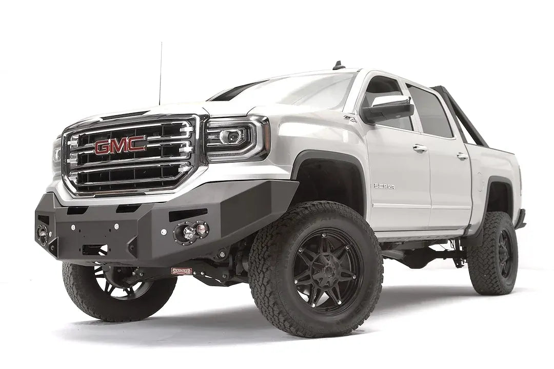 Fab Fours Premium Winch front bumper on a white GMC Sierra 1500, driver-side front three-quarter view, showing the smooth plate bumper face, winch fairlead opening and round fog lights