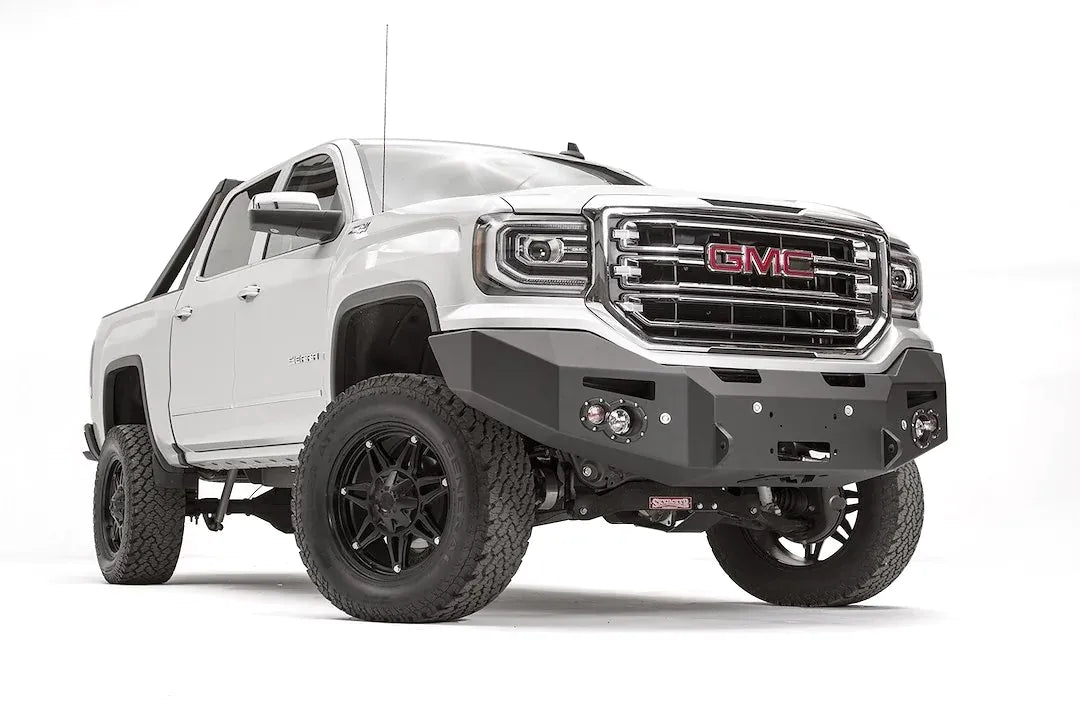 Fab Fours Premium Winch front bumper on a white GMC Sierra 1500, passenger-side front three-quarter view, showing the smooth plate bumper face, winch fairlead opening and round fog lights