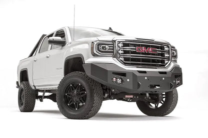 Fab Fours Premium Winch front bumper on a white GMC Sierra 1500, passenger-side front three-quarter view, showing the smooth plate bumper face, winch fairlead opening and round fog lights