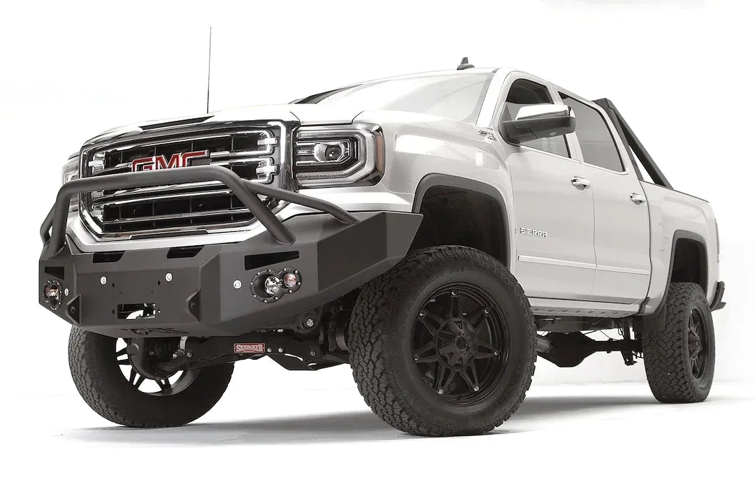 Fab Fours Premium Winch front bumper with pre-runner guard on a white GMC Sierra 1500, driver-side front three-quarter view, showing the pre-runner hoop, winch fairlead opening and round fog lights