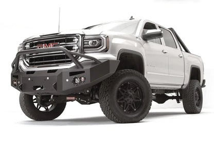 Fab Fours Premium Winch front bumper with pre-runner guard on a white GMC Sierra 1500, driver-side front three-quarter view, showing the pre-runner hoop, winch fairlead opening and round fog lights