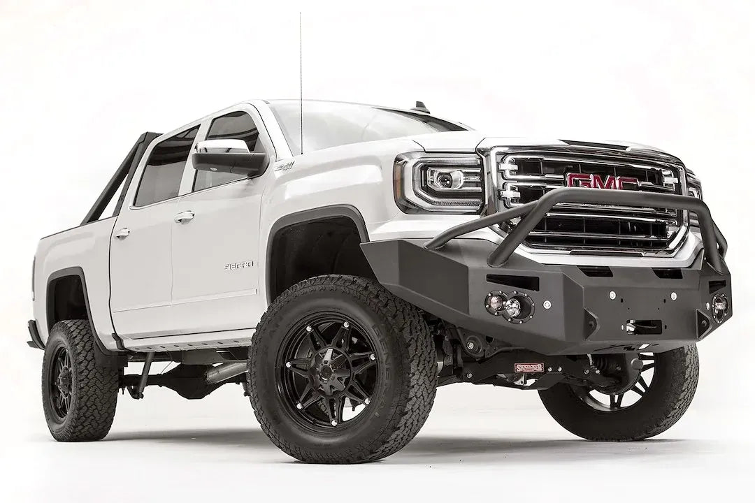 Fab Fours Premium Winch front bumper with pre-runner guard on a white GMC Sierra 1500, passenger-side front three-quarter view, showing the pre-runner hoop, winch fairlead opening and round fog lights