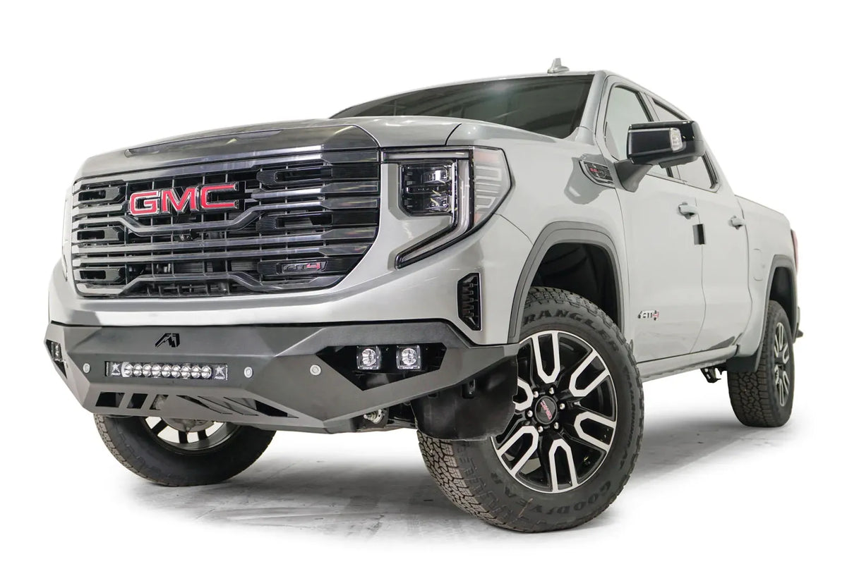 Fab Fours Vengeance front bumper on a silver GMC Sierra 1500, driver-side front three-quarter view, showing the angular bumper face, pod light cutouts and lower skid plate