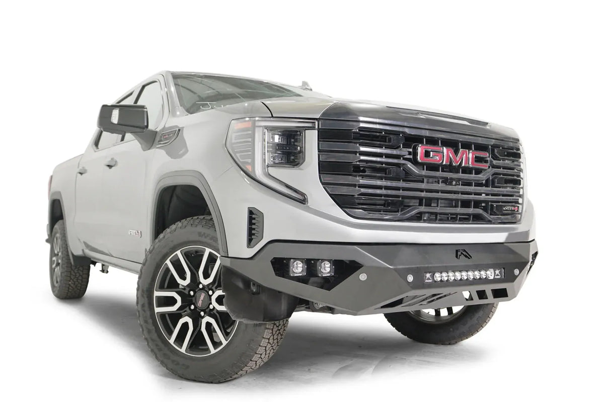 Fab Fours Vengeance front bumper on a silver GMC Sierra 1500, passenger-side front three-quarter view, showing the angular bumper face, pod light cutouts and center light bar