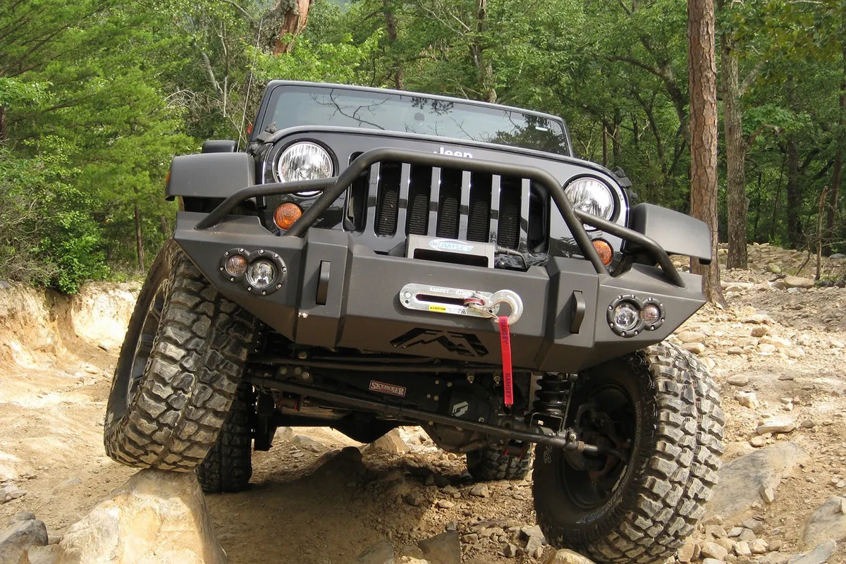 Fab Fours Lifestyle Winch bumper with pre-runner guard on a black Jeep Wrangler JK, straight-on front view outdoors on rocks, showing the pre-runner hoop, winch fairlead and round fog lights
