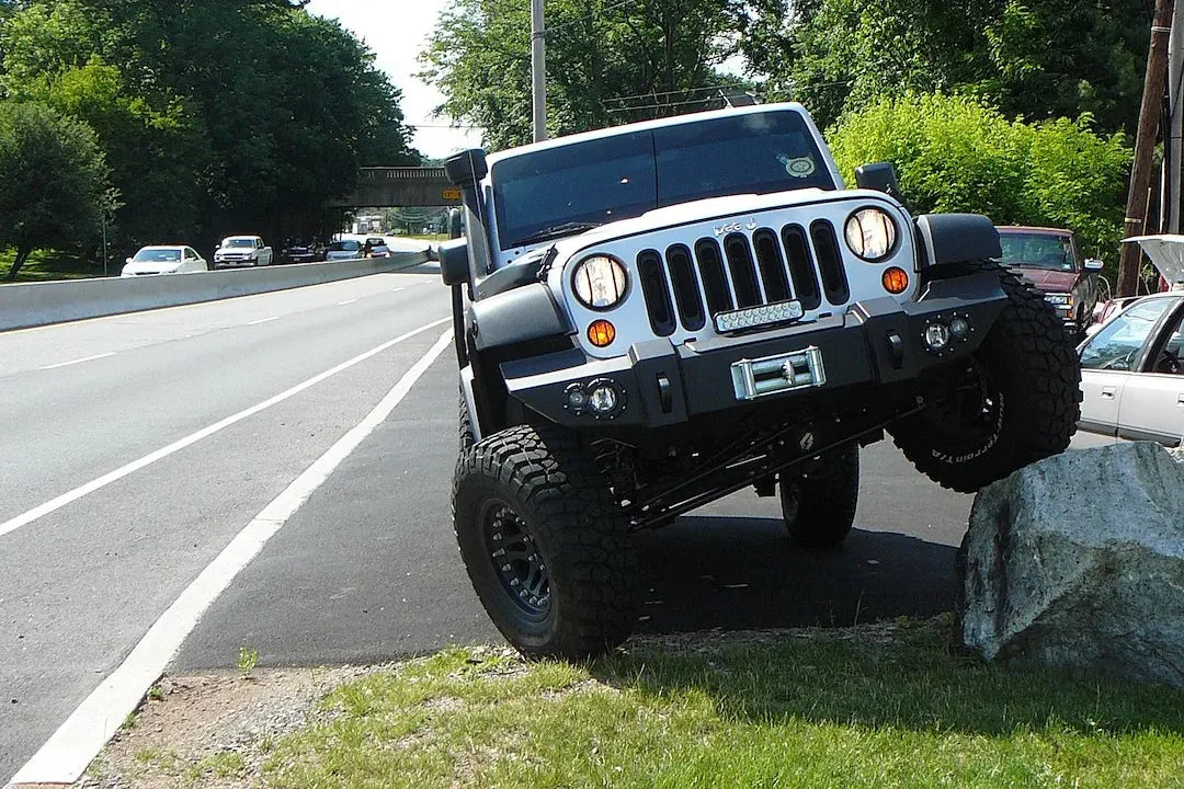 Fab Fours Lifestyle Winch bumper on a silver Jeep Wrangler JK, front view outdoors with one wheel on a boulder, showing the bumper face, winch mount and fog light openings