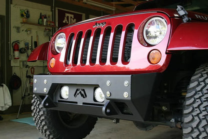 Fab Fours Full Metal Jacket stubby front bumper on a red Jeep Wrangler JK, close-up front three-quarter view in a garage, showing the bolted face plate, round fog lights and Fab Fours logo cutout