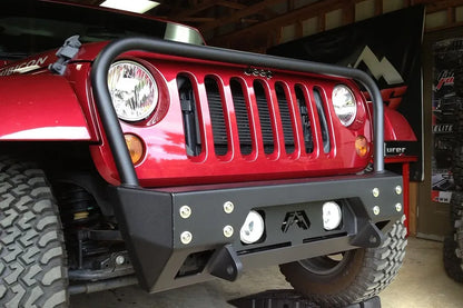 Fab Fours Full Metal Jacket stubby front bumper with full guard on a red Jeep Wrangler JK, close-up front three-quarter view in a garage, showing the tubular grille hoop, bolted face plate and round fog lights