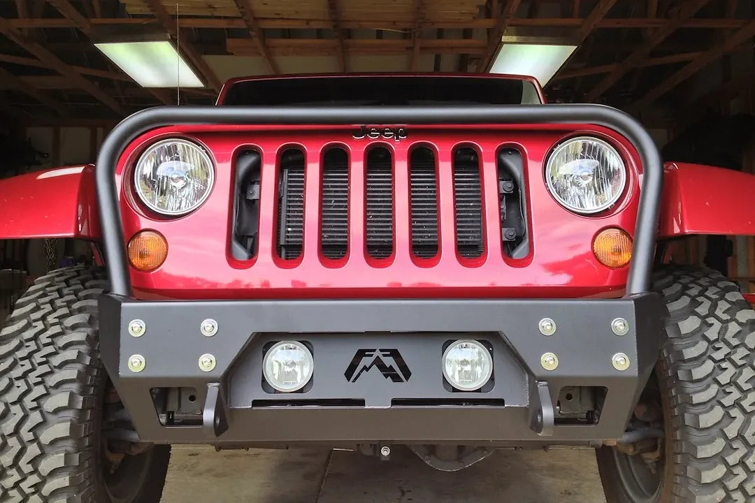 Fab Fours Full Metal Jacket stubby front bumper with full guard on a red Jeep Wrangler JK, straight-on front view in a garage, showing the tubular grille hoop, bolted face plate and round fog lights