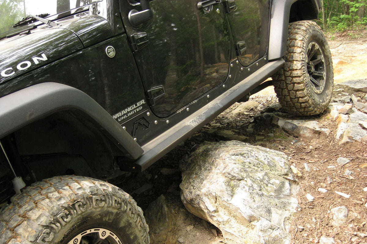 Fab Fours HardCore Rock Sliders on a black Jeep Wrangler JK Unlimited, close-up outdoors on rocks, showing the slider rail along the full rocker and riveted rocker plate