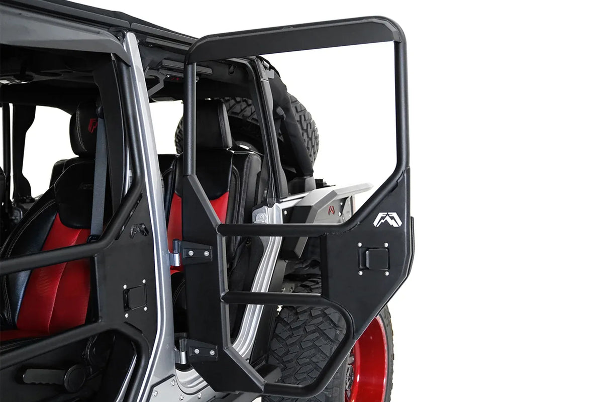 Fab Fours full tube door on a silver Jeep Wrangler JK Unlimited, driver-side rear door swung open, showing the black tubular frame with steel lower panel, paddle latch and Fab Fours logo, with the spare tire behind