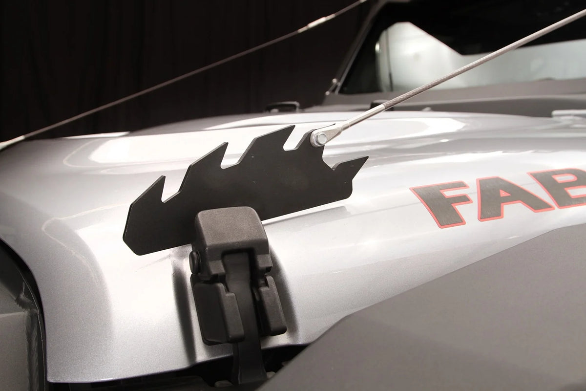 Fab Fours limb riser on a silver Jeep Wrangler, close-up of the black steel bracket with its sawtooth profile, mounted over the factory hood latch, with the cable attached at the rear tooth