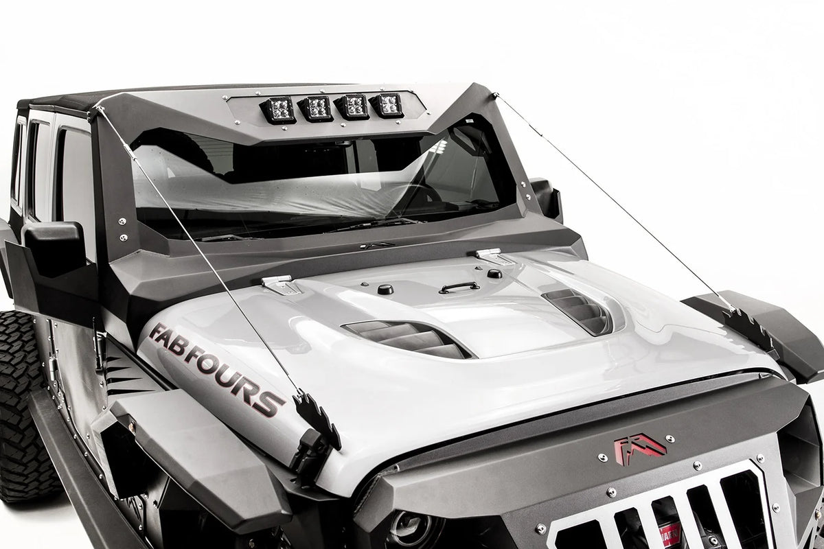 Fab Fours limb risers on a silver Jeep Wrangler, elevated front view, showing steel cables running from the jagged hood-mounted brackets to the corners of the Fab Fours windshield frame with pod lights