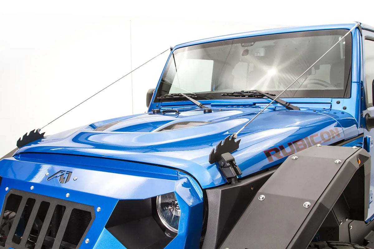 Fab Fours limb riser on a blue Jeep Wrangler JK Rubicon, elevated front view showing both stainless cables stretched from the hood brackets to the top corners of the windshield