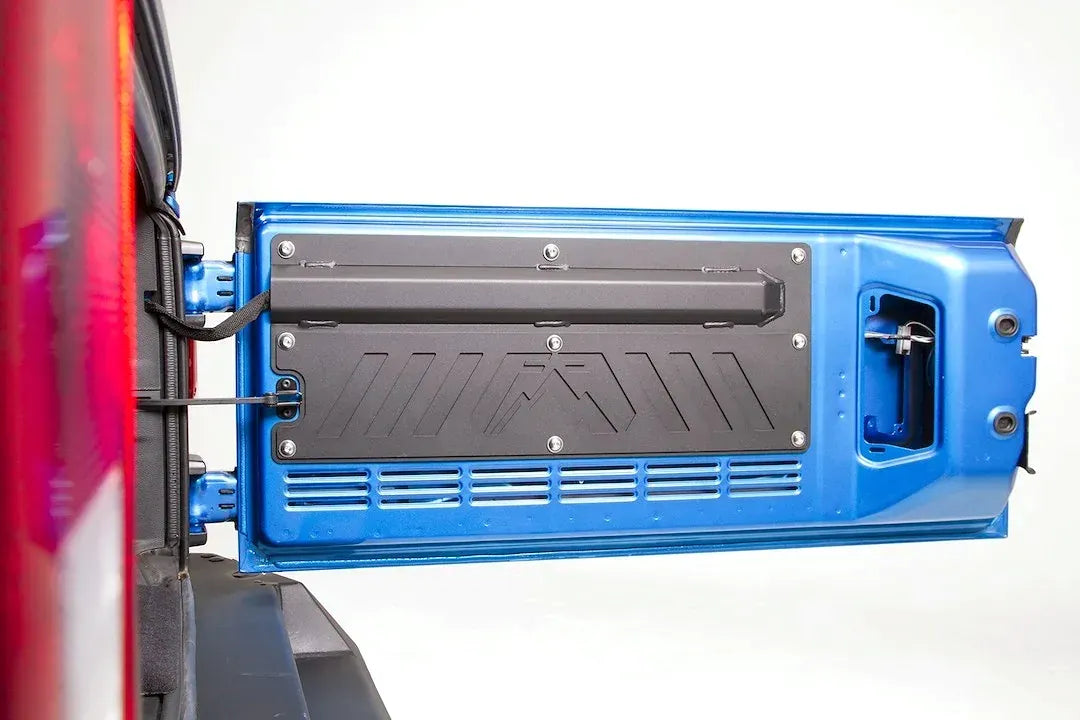 Fab Fours tailgate plate on the inside of a blue Jeep Wrangler JK tailgate, straight-on close-up, showing the integrated storage tube, the stamped Fab Fours logo and the bolt-on mounting