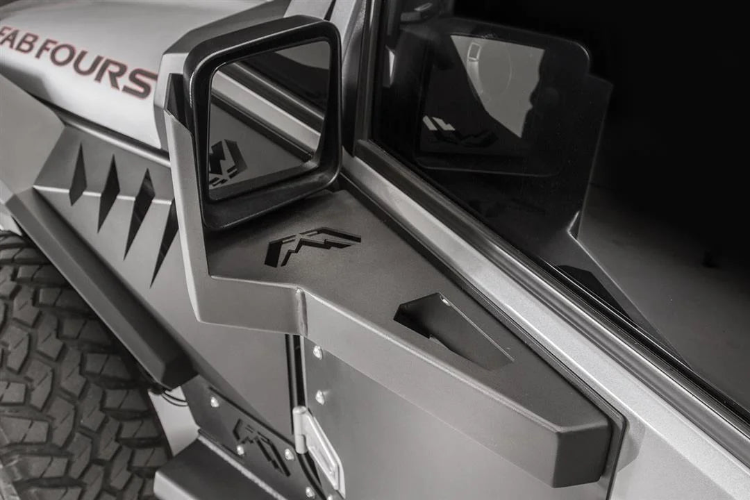 Fab Fours door skin mirror guard on a gray Jeep Wrangler JK, driver side, showing the angular steel mirror mount with Fab Fours logo cutout beside the hood and fender armor