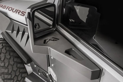 Fab Fours door skin mirror guard on a gray Jeep Wrangler JK, driver side, showing the angular steel mirror mount with Fab Fours logo cutout beside the hood and fender armor