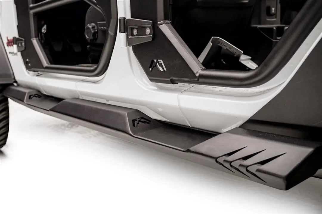 Fab Fours Light Rock Slider on a white Jeep Wrangler JL, low driver-side view, showing the flat black rocker guard with Fab Fours logo cutouts and chevron-cut rear end