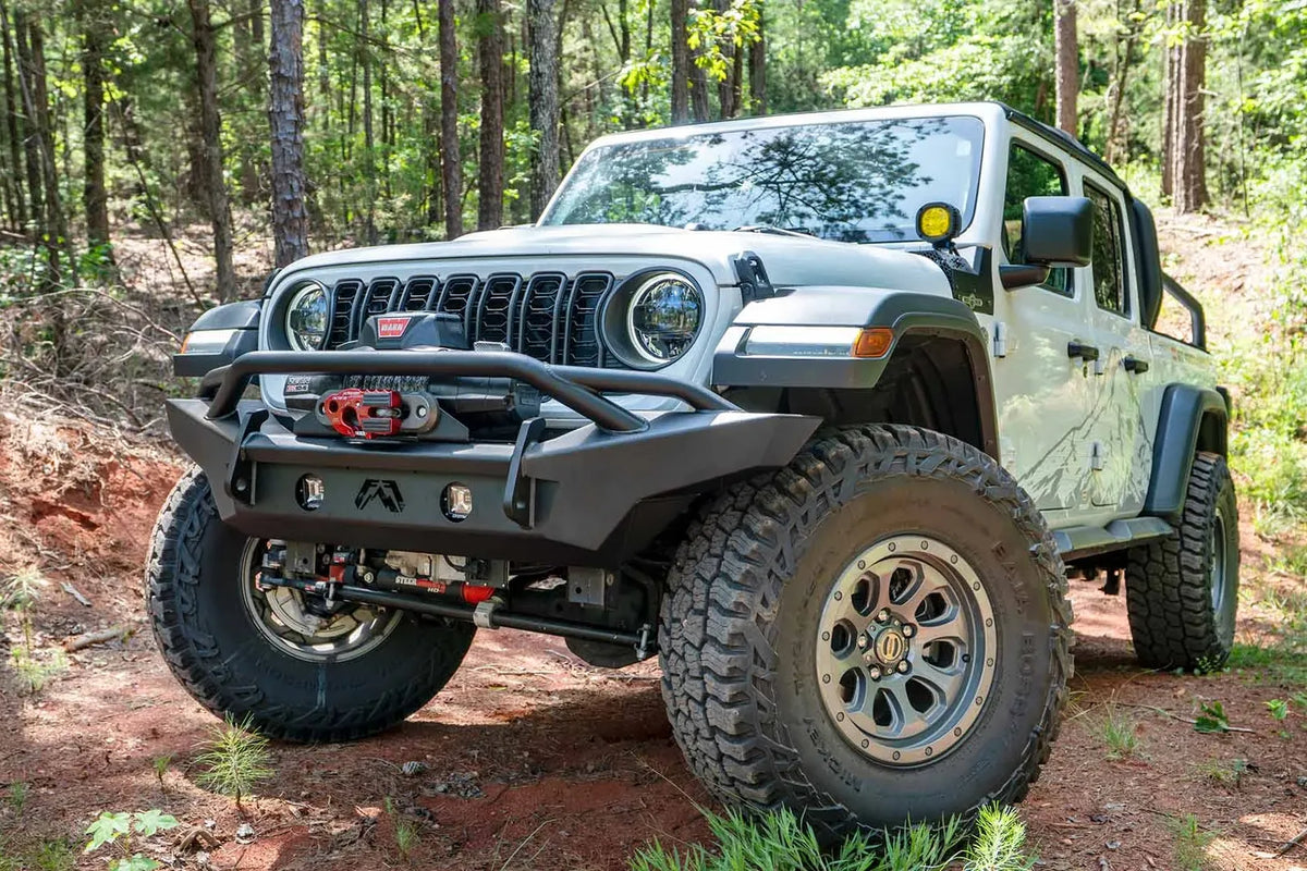 Fab Fours Premium front Lifestyle bumper with pre-runner guard on a white Jeep Wrangler JL on a dirt trail, driver-side front three-quarter view, showing the tube hoop, winch with red hook and round lights