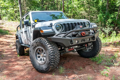 Fab Fours Premium front Lifestyle bumper with pre-runner guard on a white Jeep Wrangler JL in the woods, passenger-side front three-quarter view, showing the tube hoop, winch and round lights