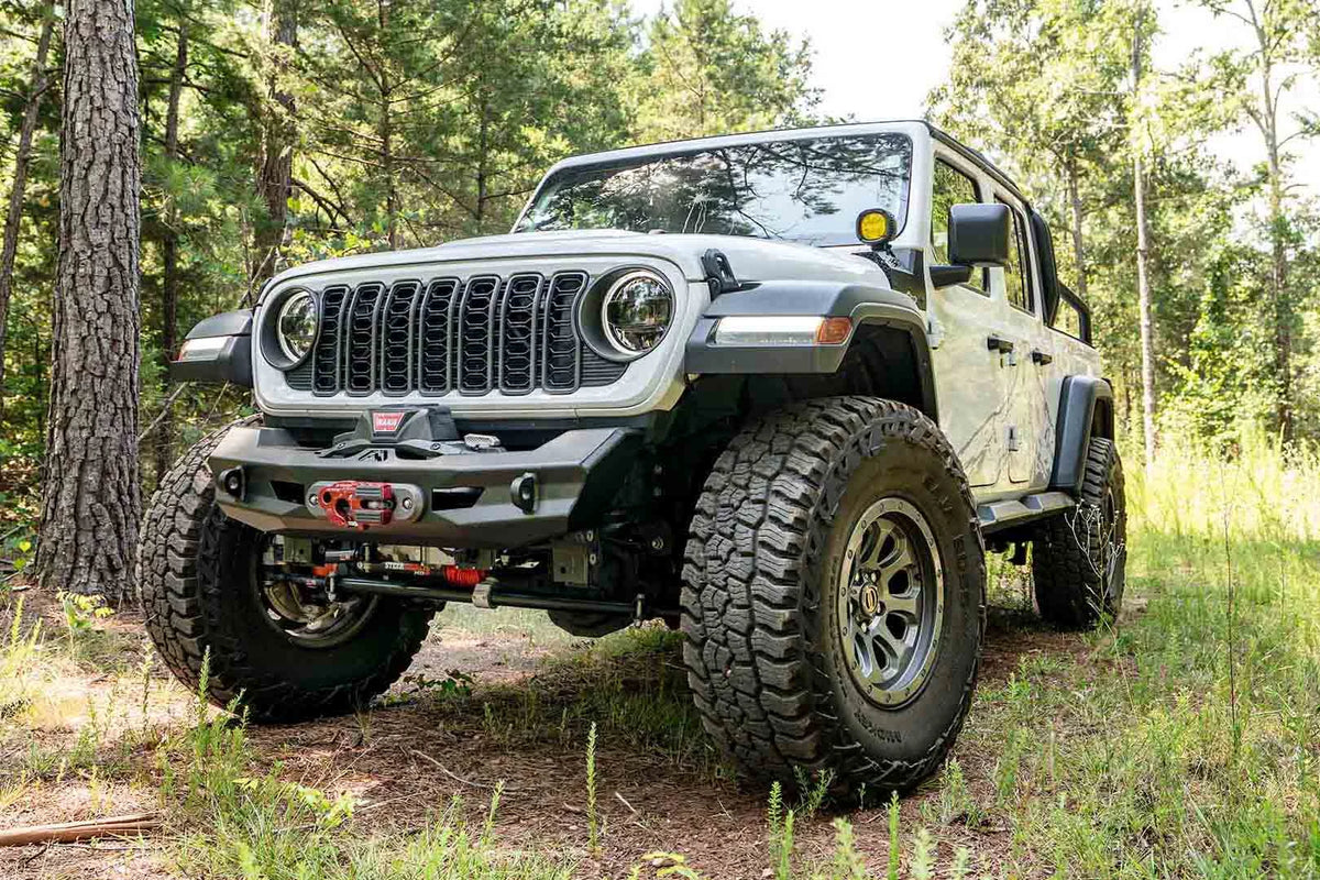 Fab Fours Ghost stubby front winch bumper with no guard on a lifted white Jeep Wrangler JL on a wooded trail, front driver-side view, showing the compact bumper with winch, fairlead and D-ring mounts