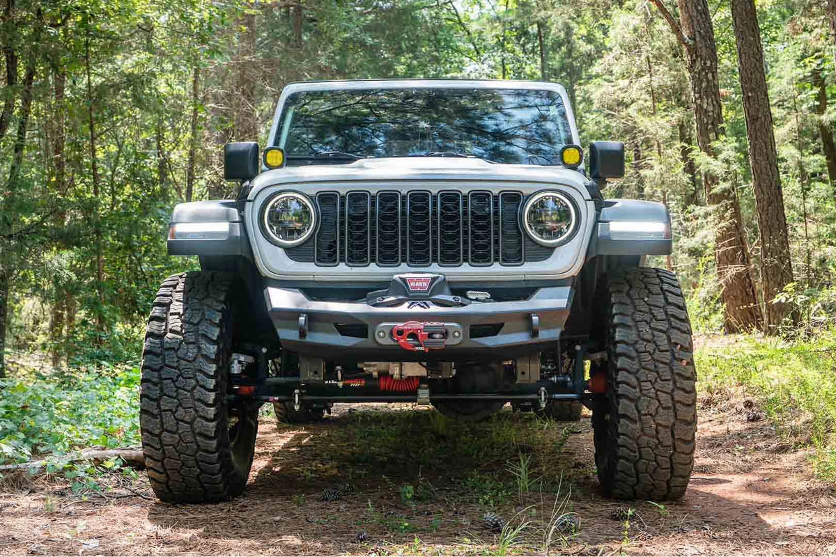 Fab Fours Ghost stubby front winch bumper with no guard on a white Jeep Wrangler JL on a wooded trail, straight-on front view, showing the stubby bumper with a centered winch and fairlead