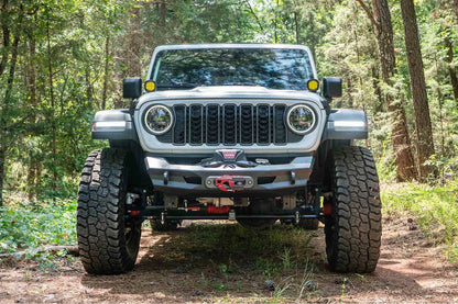 Fab Fours Ghost stubby front winch bumper with no guard on a white Jeep Wrangler JL on a wooded trail, straight-on front view, showing the stubby bumper with a centered winch and fairlead