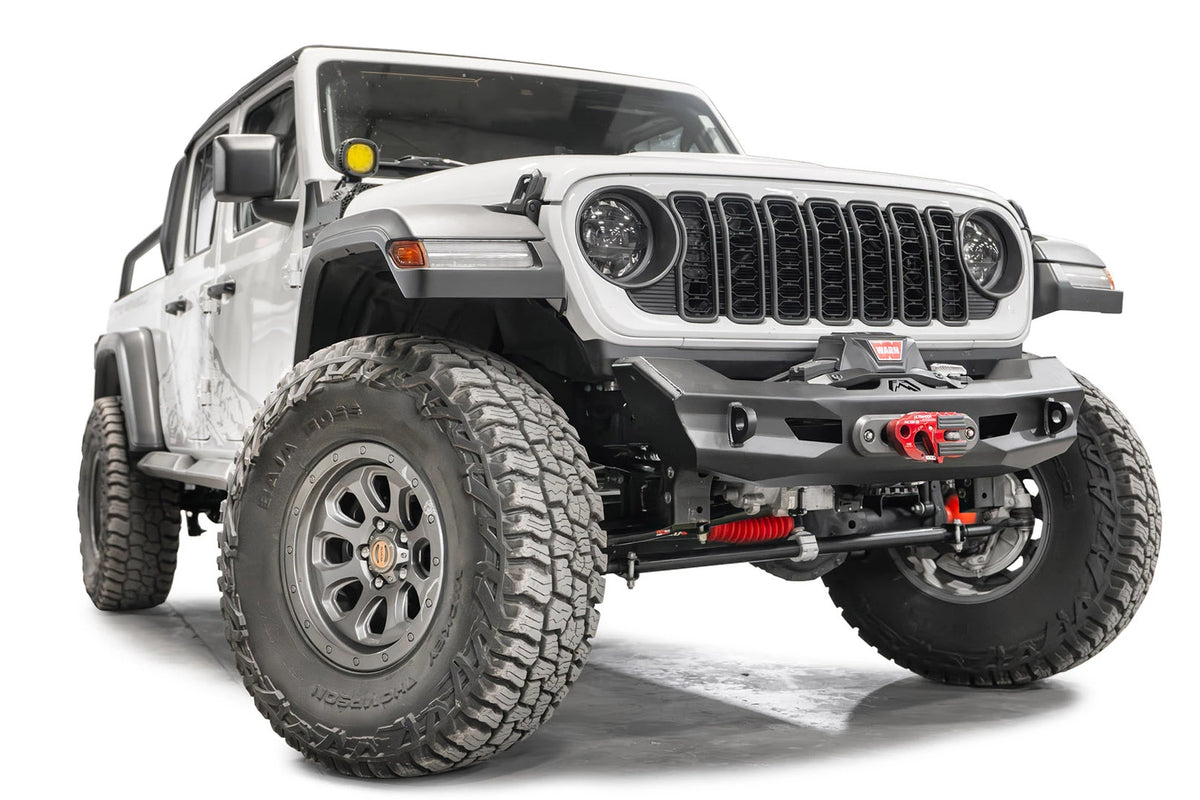 Fab Fours Ghost stubby front winch bumper with no guard on a lifted white Jeep Wrangler JL, front passenger-side view, showing the stubby bumper ends that clear the tires, the winch fairlead and the D-ring mounts