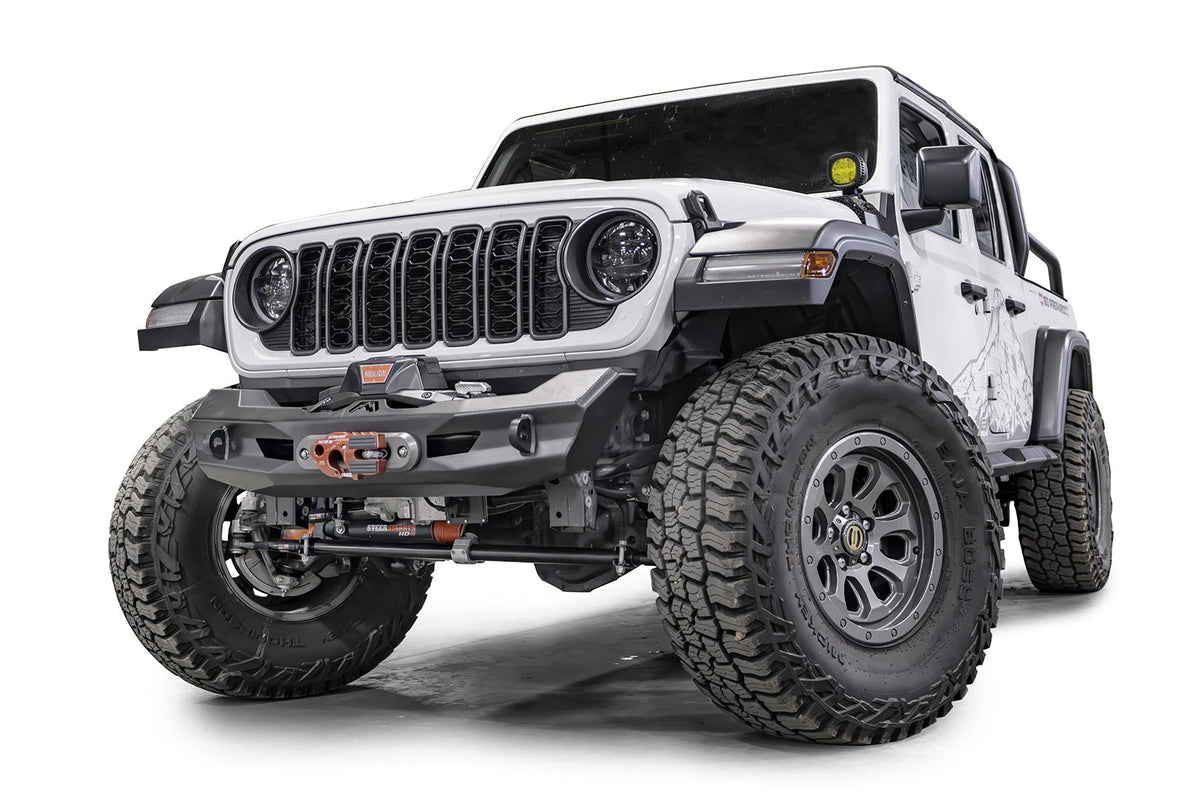 Fab Fours Ghost stubby front winch bumper with no guard on a lifted white Jeep Wrangler JL, front driver-side view, showing the compact faceted steel bumper with a centered winch and fairlead, Fab Fours logo and D-ring mounts