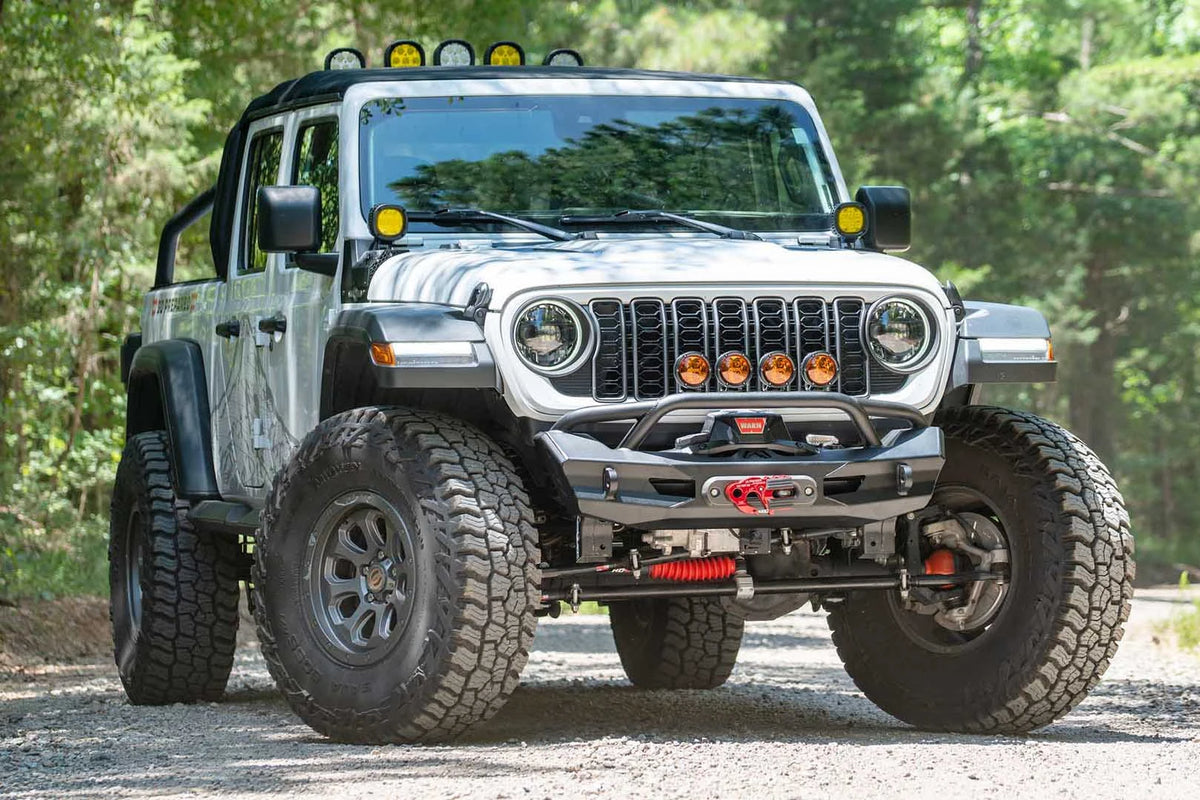 Fab Fours Ghost stubby front winch bumper with pre-runner guard on a lifted white Jeep Wrangler JL on a gravel trail, front passenger-side view, showing the tubular pre-runner hoop, winch, fairlead and the stubby bumper ends clearing the tires