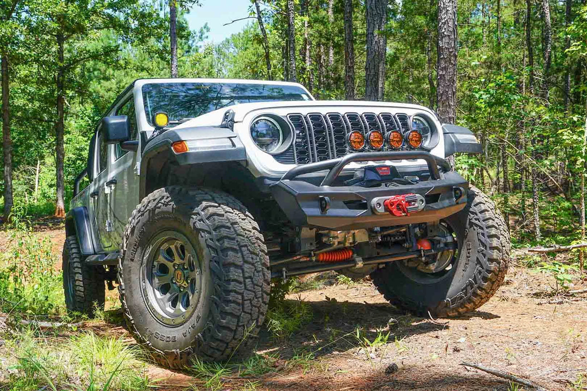 Fab Fours Ghost stubby front winch bumper with pre-runner guard on a lifted white Jeep Wrangler JL on a wooded trail, front passenger-side view, showing the pre-runner hoop, winch fairlead and D-ring mounts on the compact faceted bumper