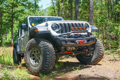Fab Fours Ghost stubby front winch bumper with pre-runner guard on a lifted white Jeep Wrangler JL on a wooded trail, front passenger-side view, showing the pre-runner hoop, winch fairlead and D-ring mounts on the compact faceted bumper