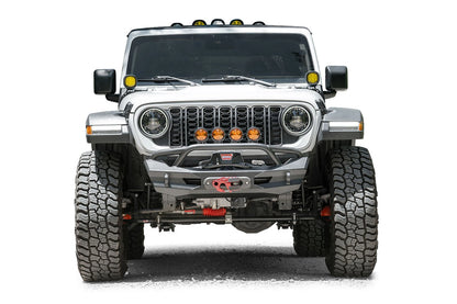 Fab Fours Ghost stubby front winch bumper with pre-runner guard on a lifted white Jeep Wrangler JL, straight-on front view, showing the tubular pre-runner hoop over a centered winch and fairlead, with four amber pod lights mounted in the grille