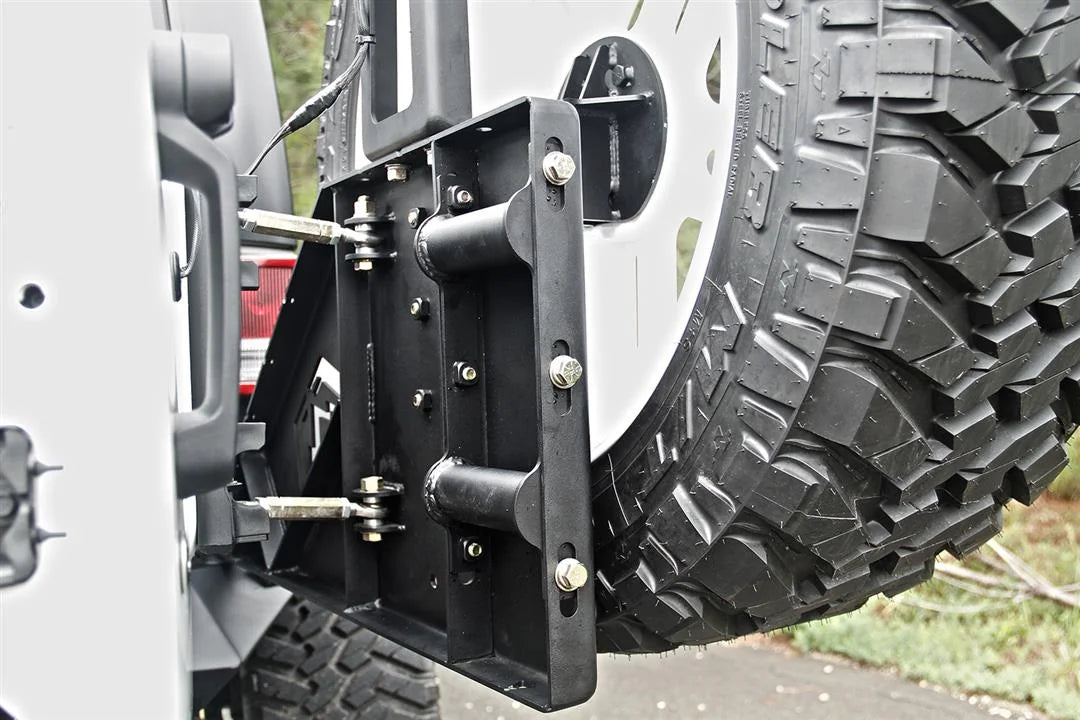 Fab Fours off-the-door tire carrier on a white Jeep Wrangler JK, outdoor close-up of the tire mount plate, showing the slotted bolt-on mounts, the linkage rods and the spare tire