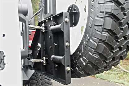 Fab Fours off-the-door tire carrier on a white Jeep Wrangler JK, outdoor close-up of the tire mount plate, showing the slotted bolt-on mounts, the linkage rods and the spare tire
