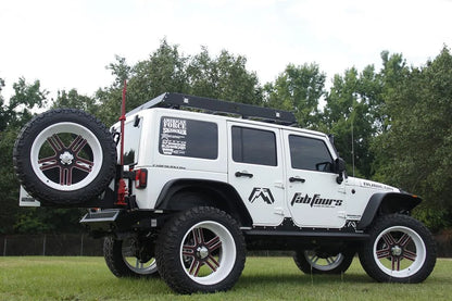 Fab Fours off-the-door tire carrier on a lifted white Jeep Wrangler JK Unlimited, rear passenger-side view, showing the spare tire and jack mount on the carrier above a Fab Fours rear bumper, with a Fab Fours roof rack